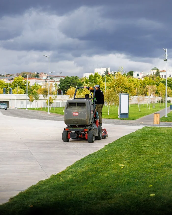 A park employee driving a lawnmower on a cloudy day in an urban park setting.